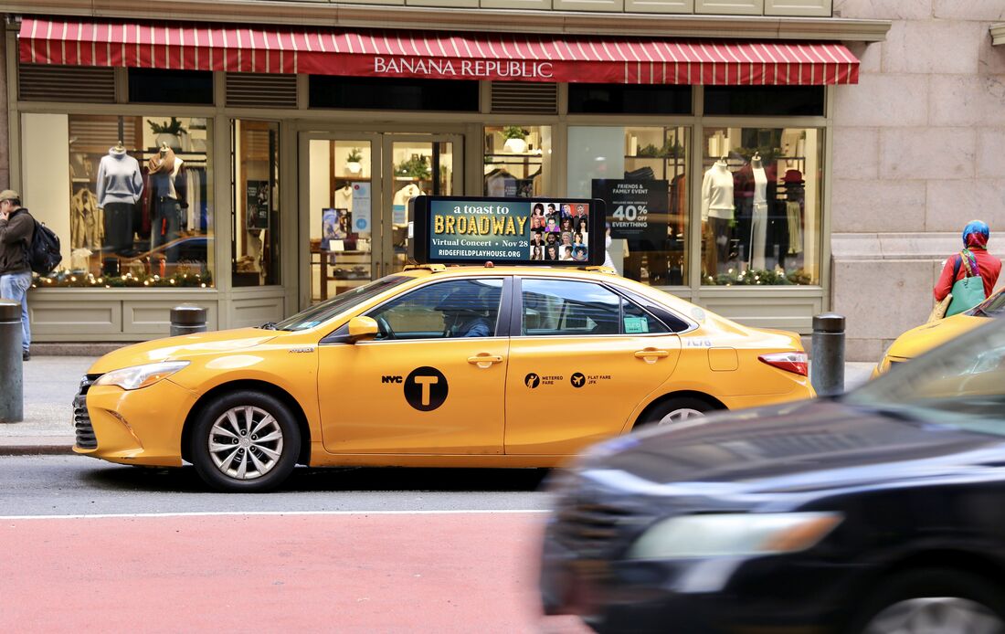 NYC yellow taxi with 'A Toast to Broadway' roof-top screen ad, storefront in background
