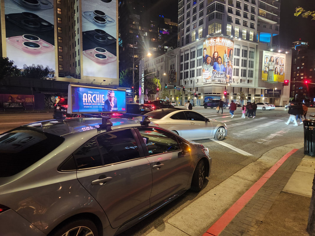 Los Angeles nighttime street with car-top screen showing BritBox 'Archie' ad