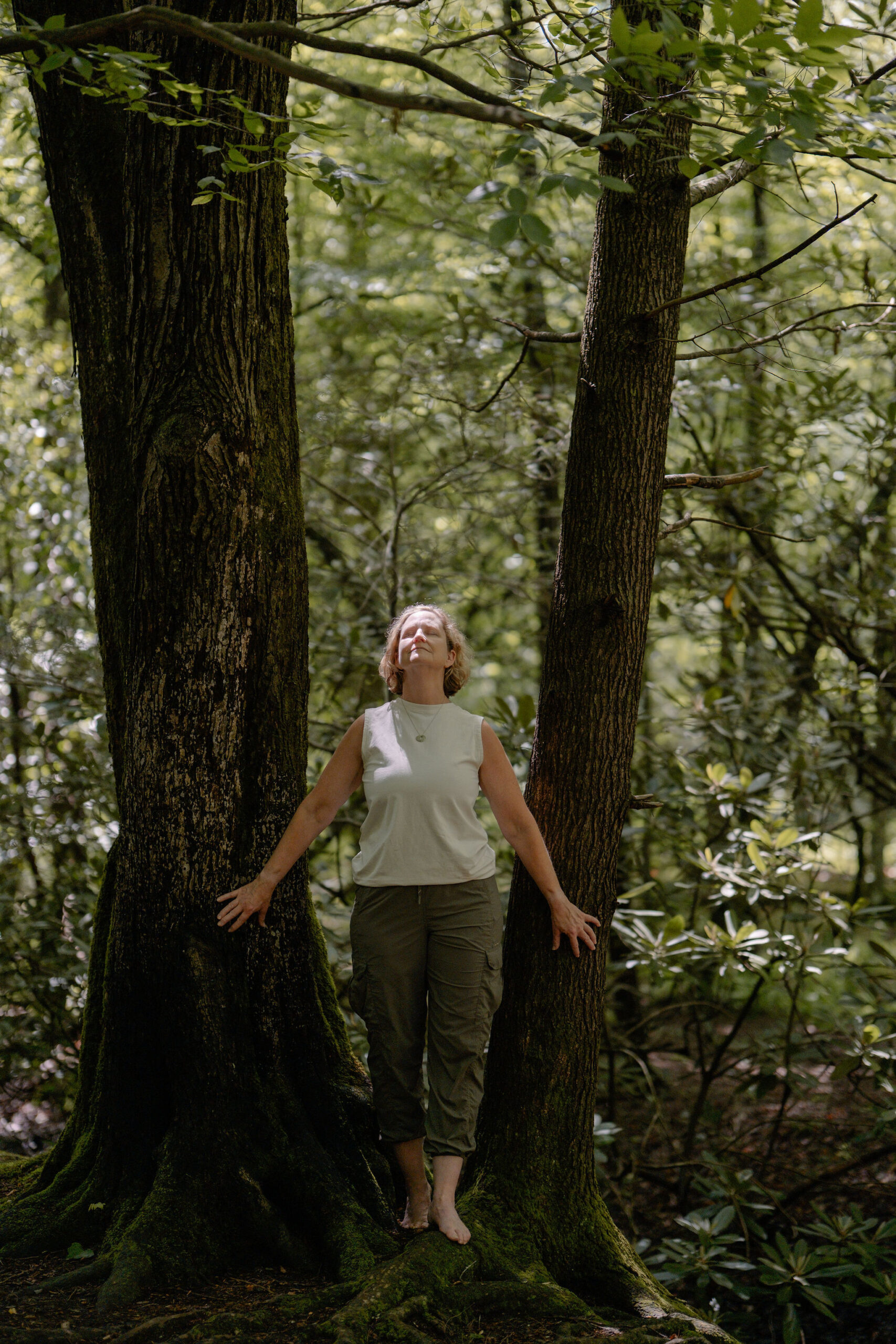 Booth Andrews standing barefoot between two large forest trees with arms outstretched
