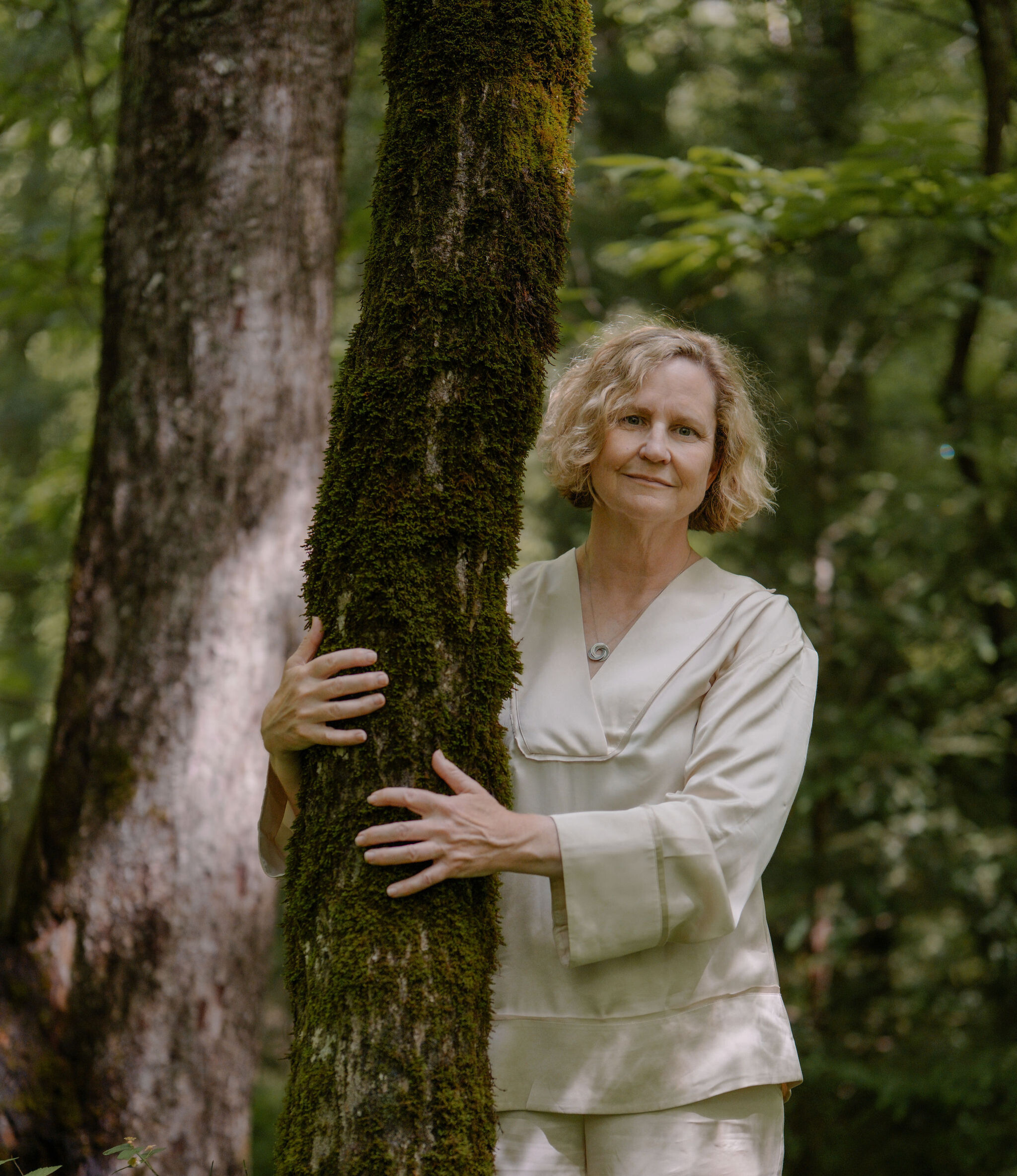 Booth Andrews in a cream outfit embracing a moss-covered tree in a forest