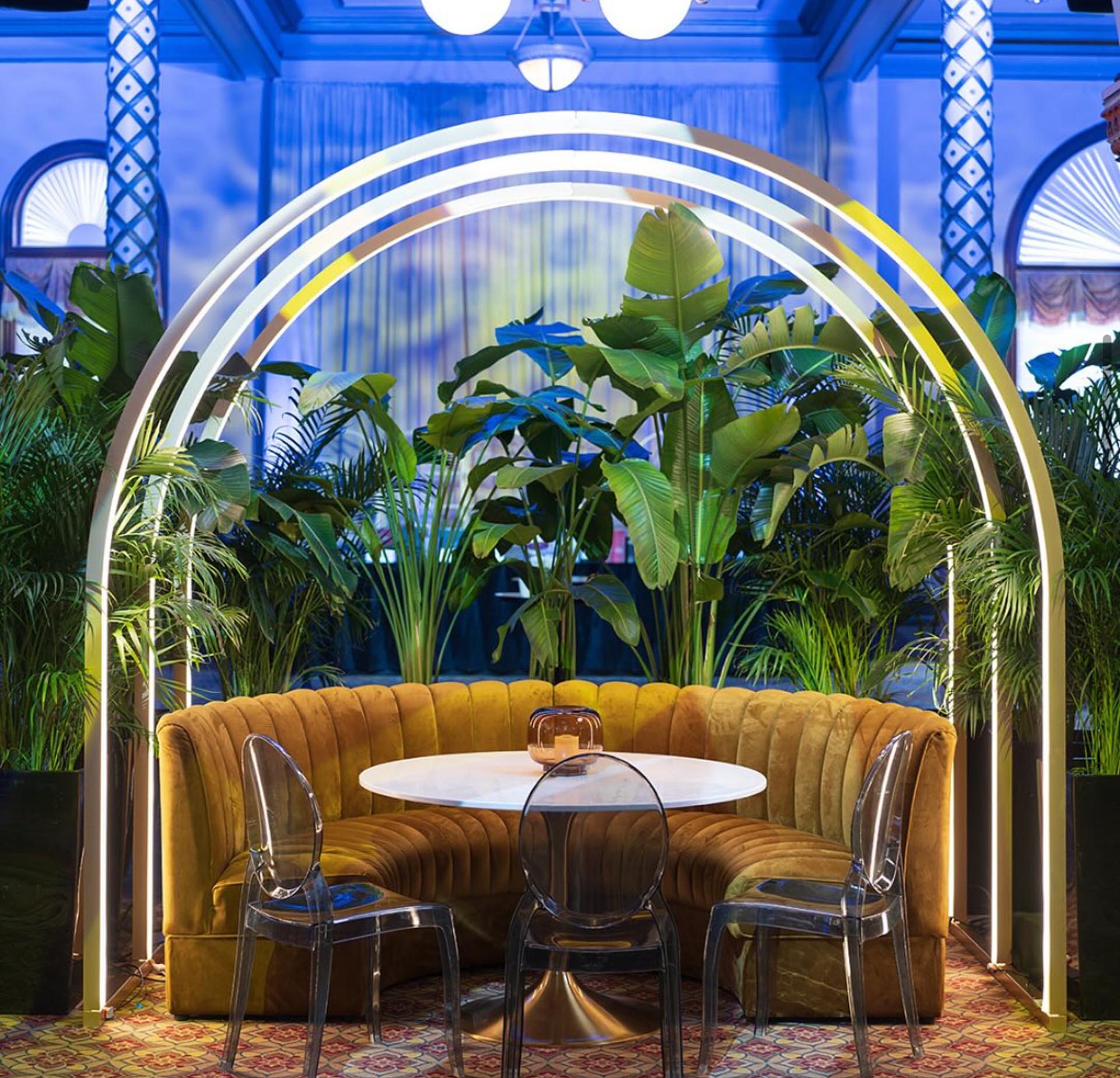 Glamorous corporate event vignette — curved mustard velvet banquette under a neon arch with tropical plants