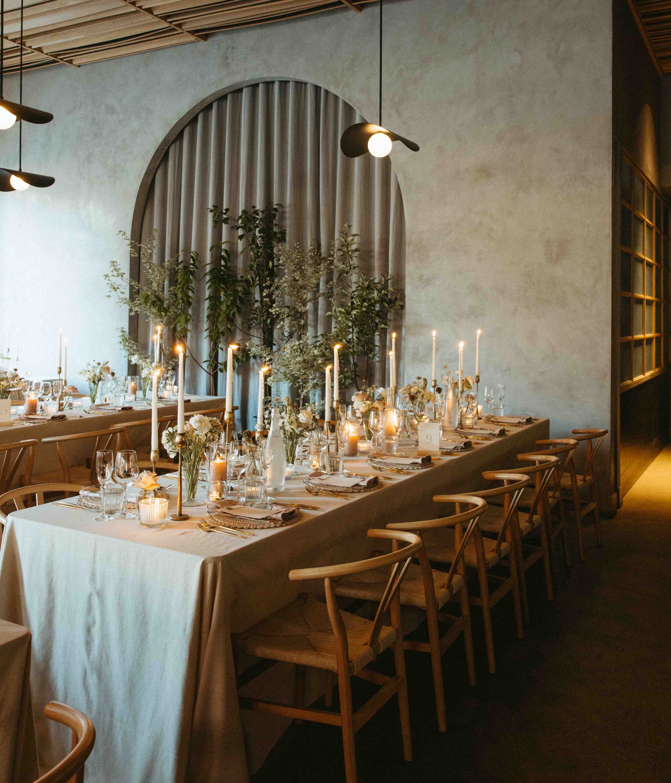 Elegant long dining table set with wishbone chairs, candles and greenery in an arched alcove