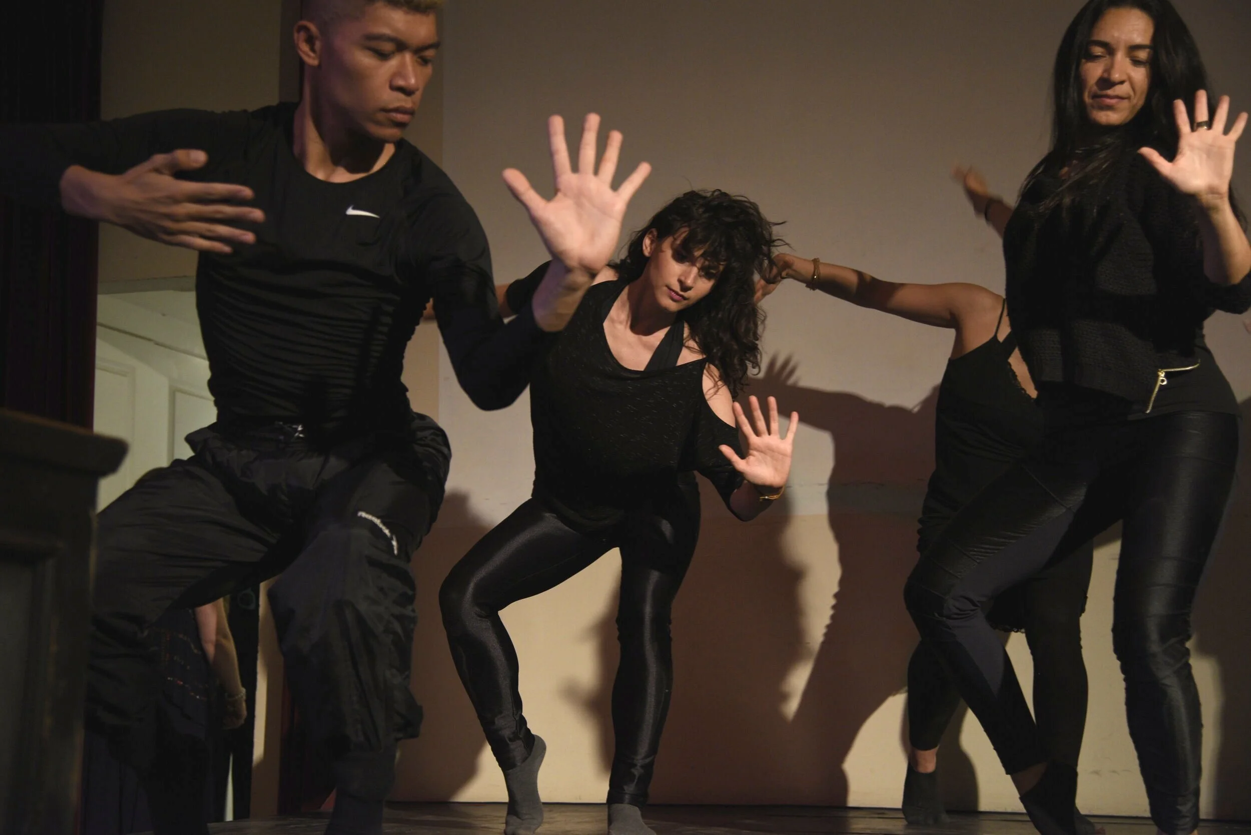 Three dancers in black moving expressively against a warm beige wall