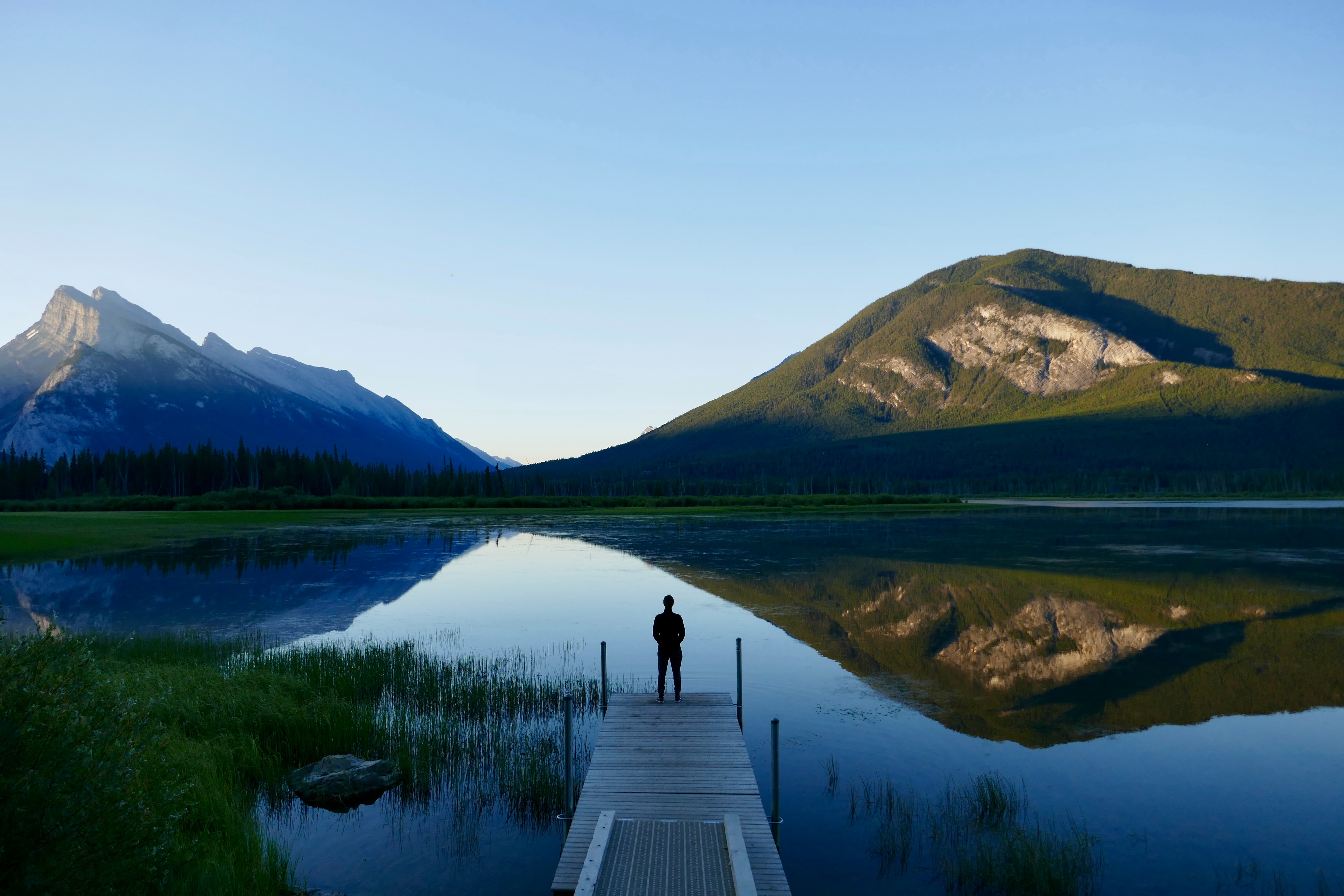 A man standing alone on a dock at dusk looking out over a mountain lake