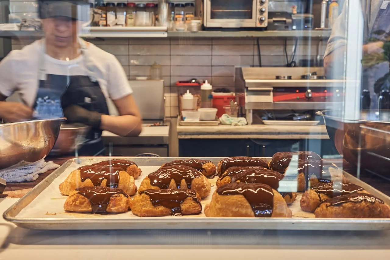 Tray of chocolate-glazed croissants with a baker at work behind the glass