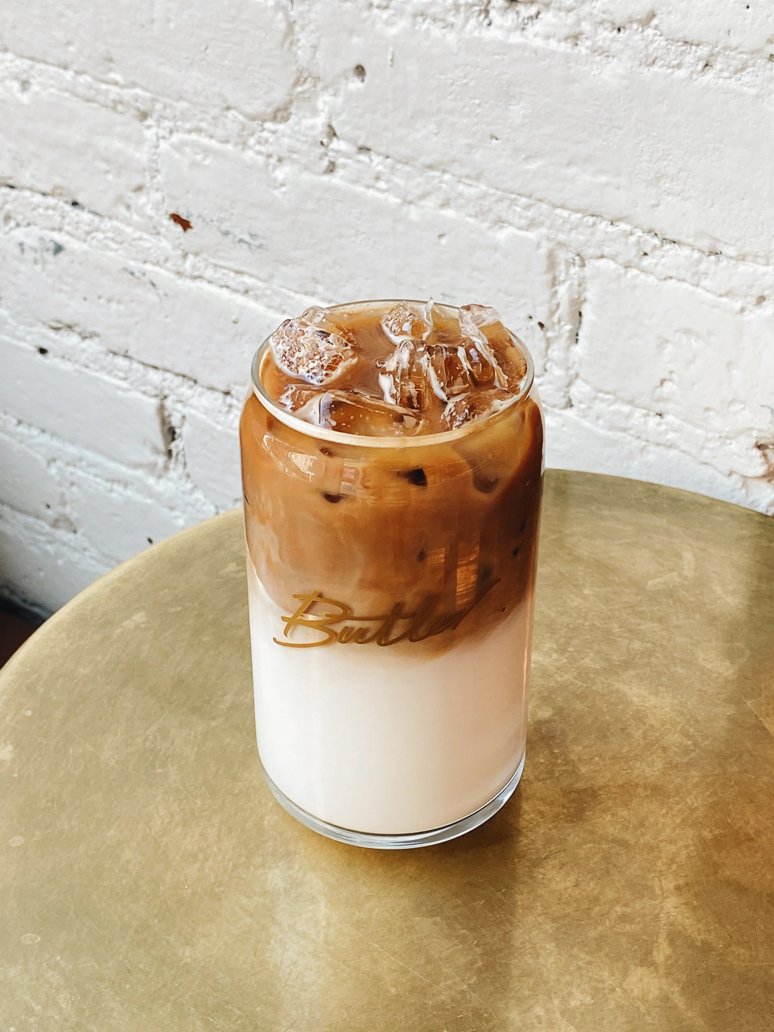 Iced coffee in a branded Butler glass can on a brass table against a white brick wall