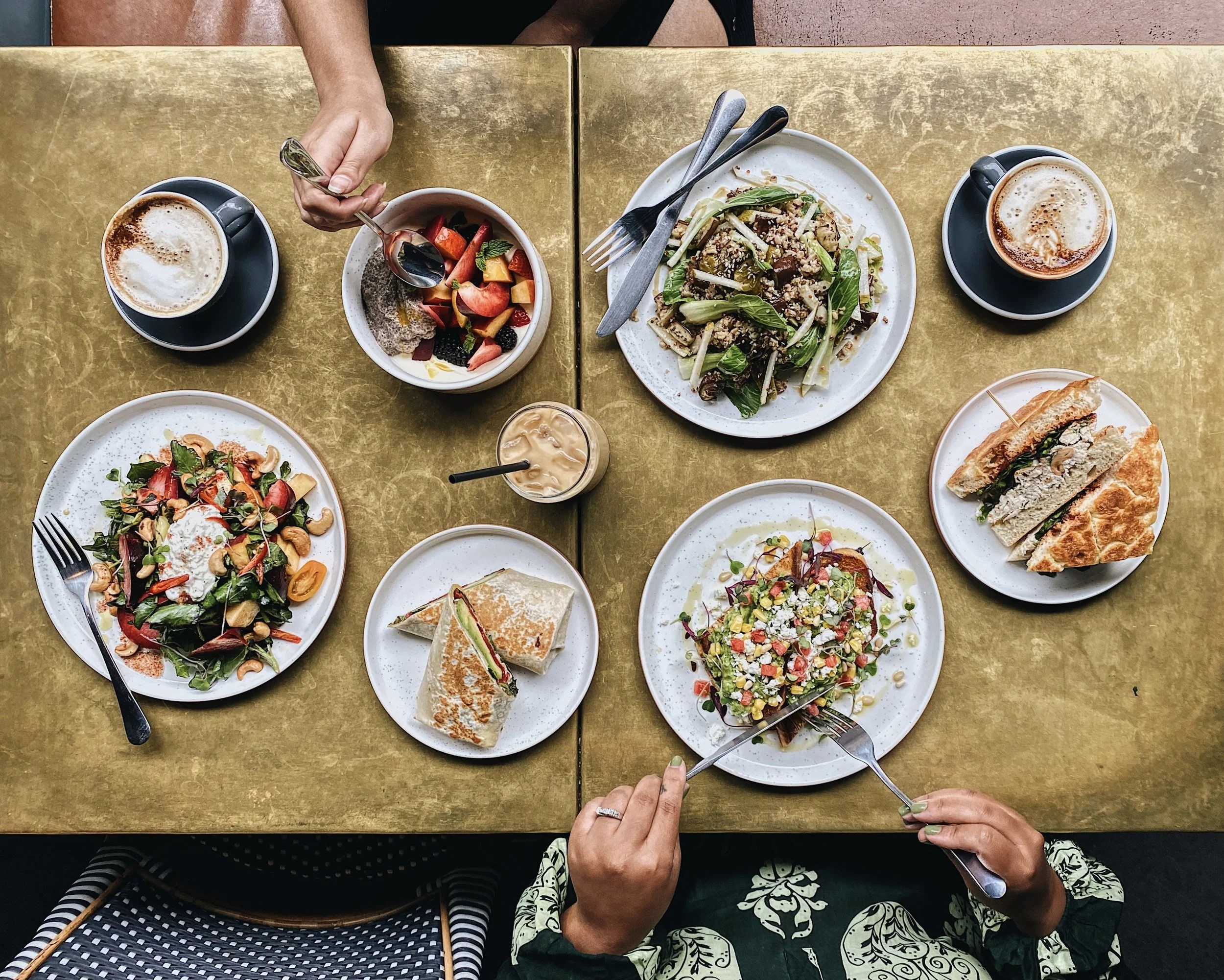 Overhead flatlay of dishes and lattes on a gold table with two diners' hands