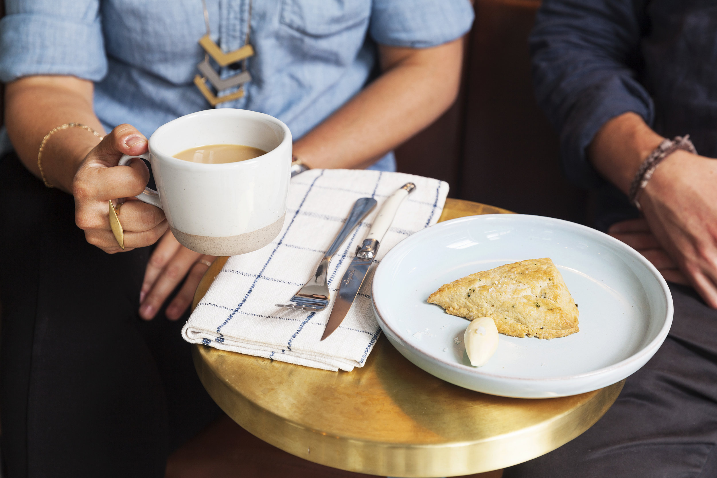 Hands around a coffee mug with a scone on a pale blue plate on a brass table