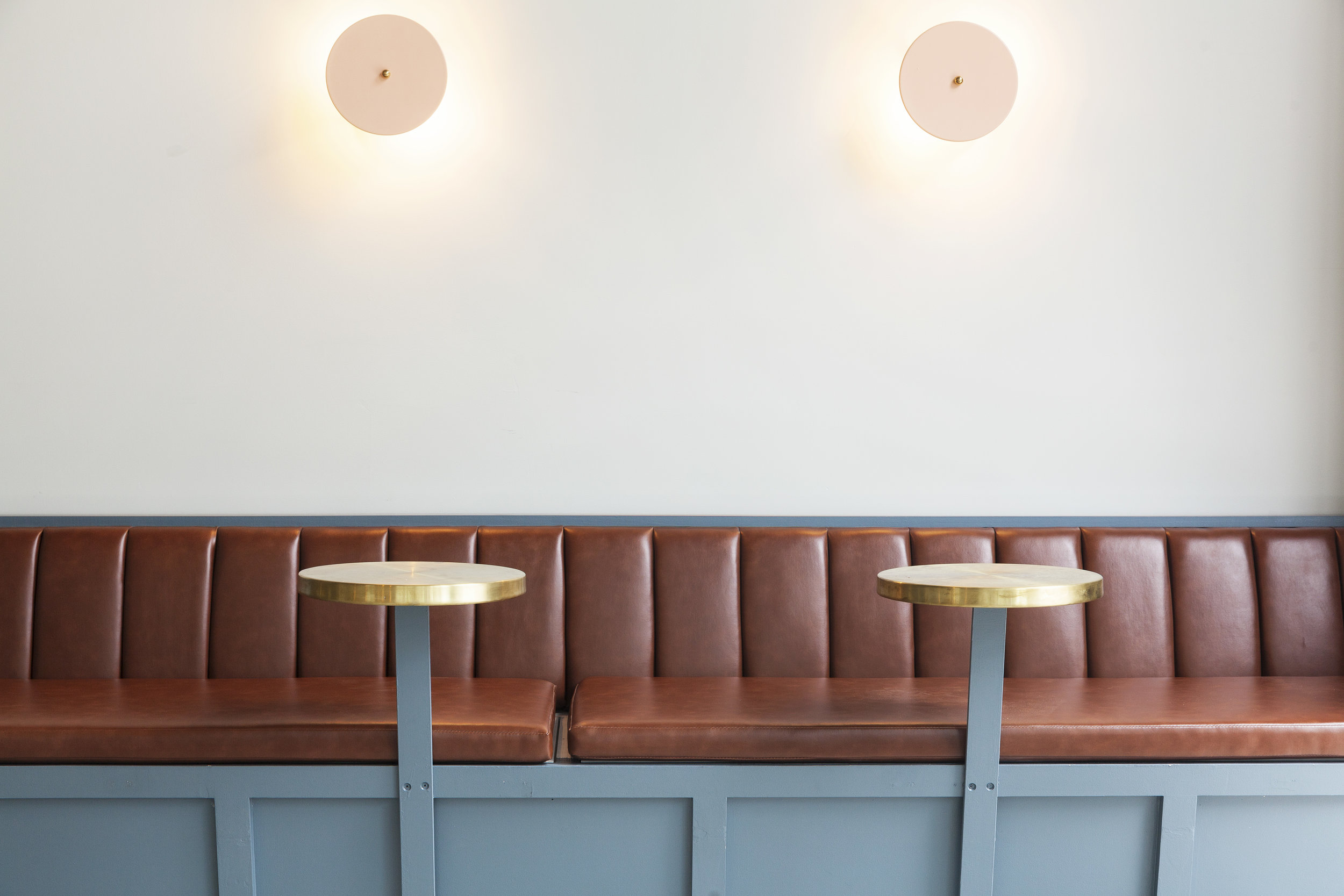 Brown leather banquette with two brass round tables and two pink disc wall sconces
