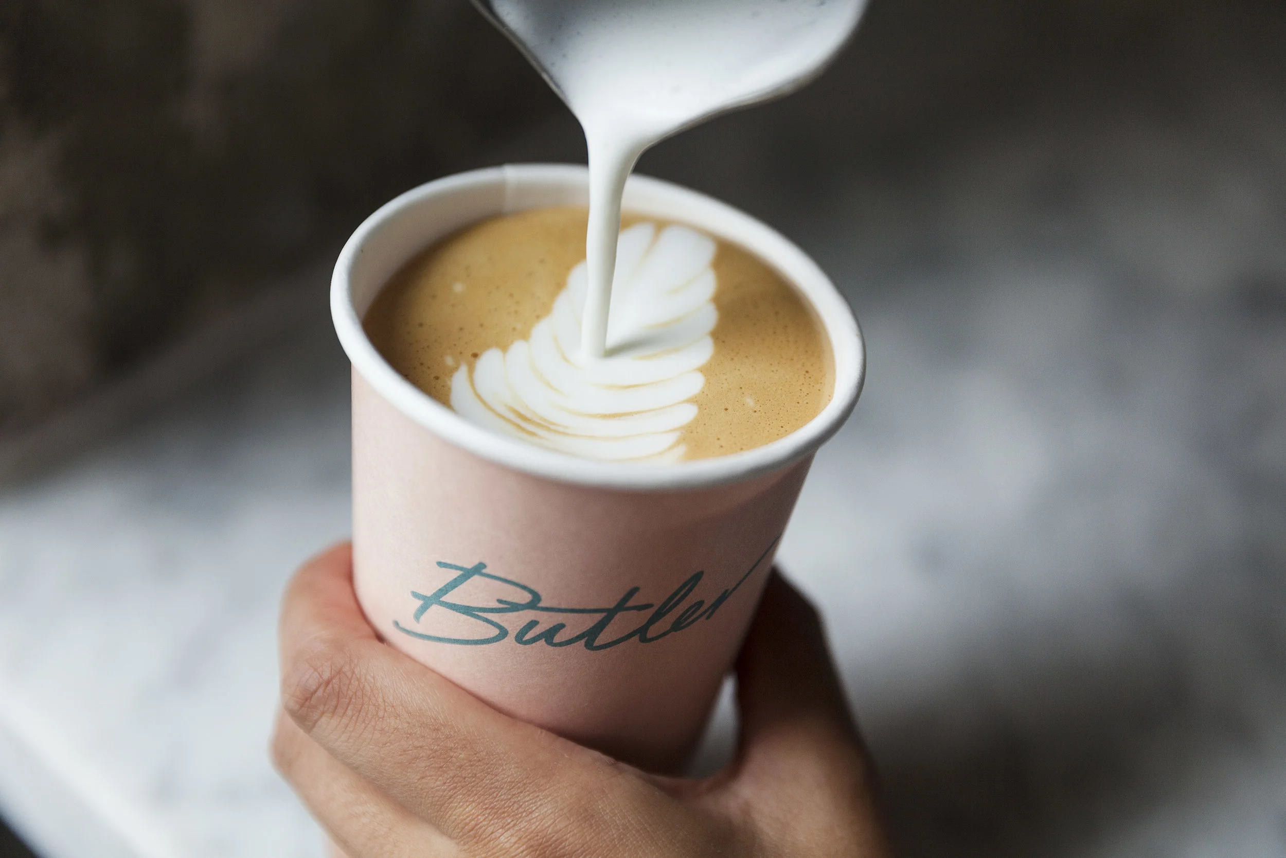 Hand holding a Butler-branded teal cup as steamed milk is poured into latte art
