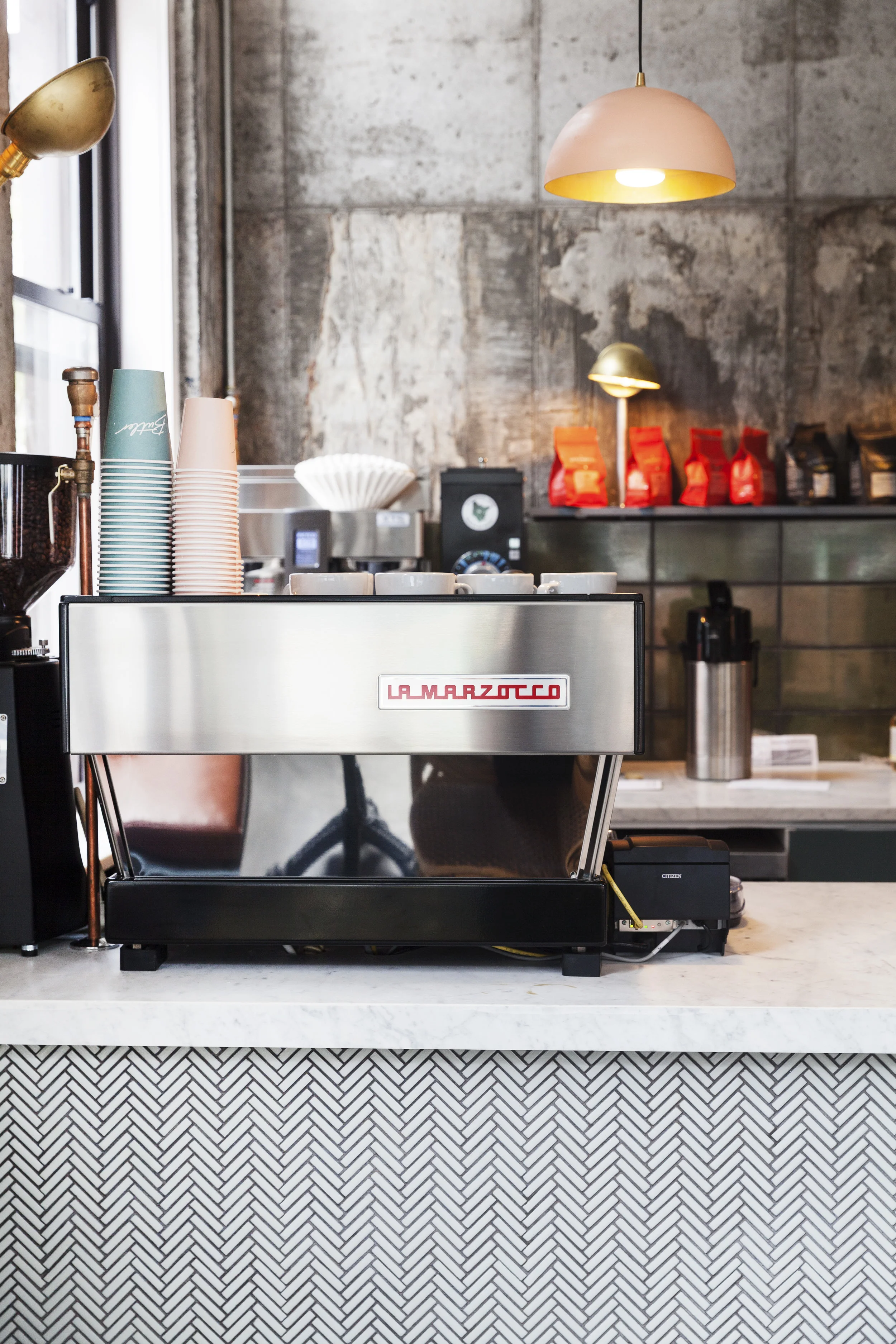 La Marzocco espresso machine on a marble herringbone counter with a pink pendant lamp and stacked cups