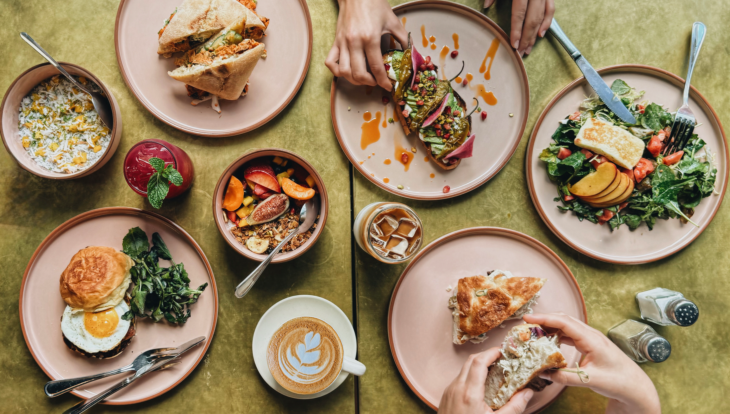 Overhead flatlay of Butler dishes — sandwiches, avocado toast, fruit bowl, salad and a latte on a gold-green table