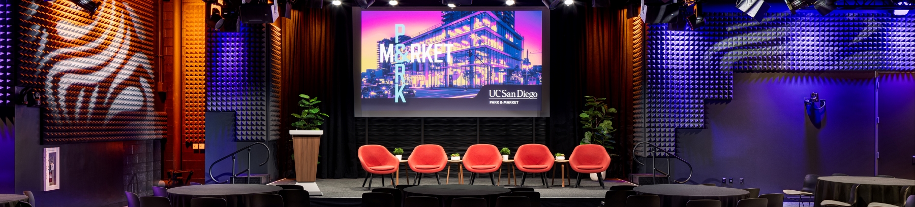 Interior of UC San Diego Park & Market event venue with stage, red chairs, and large display screen