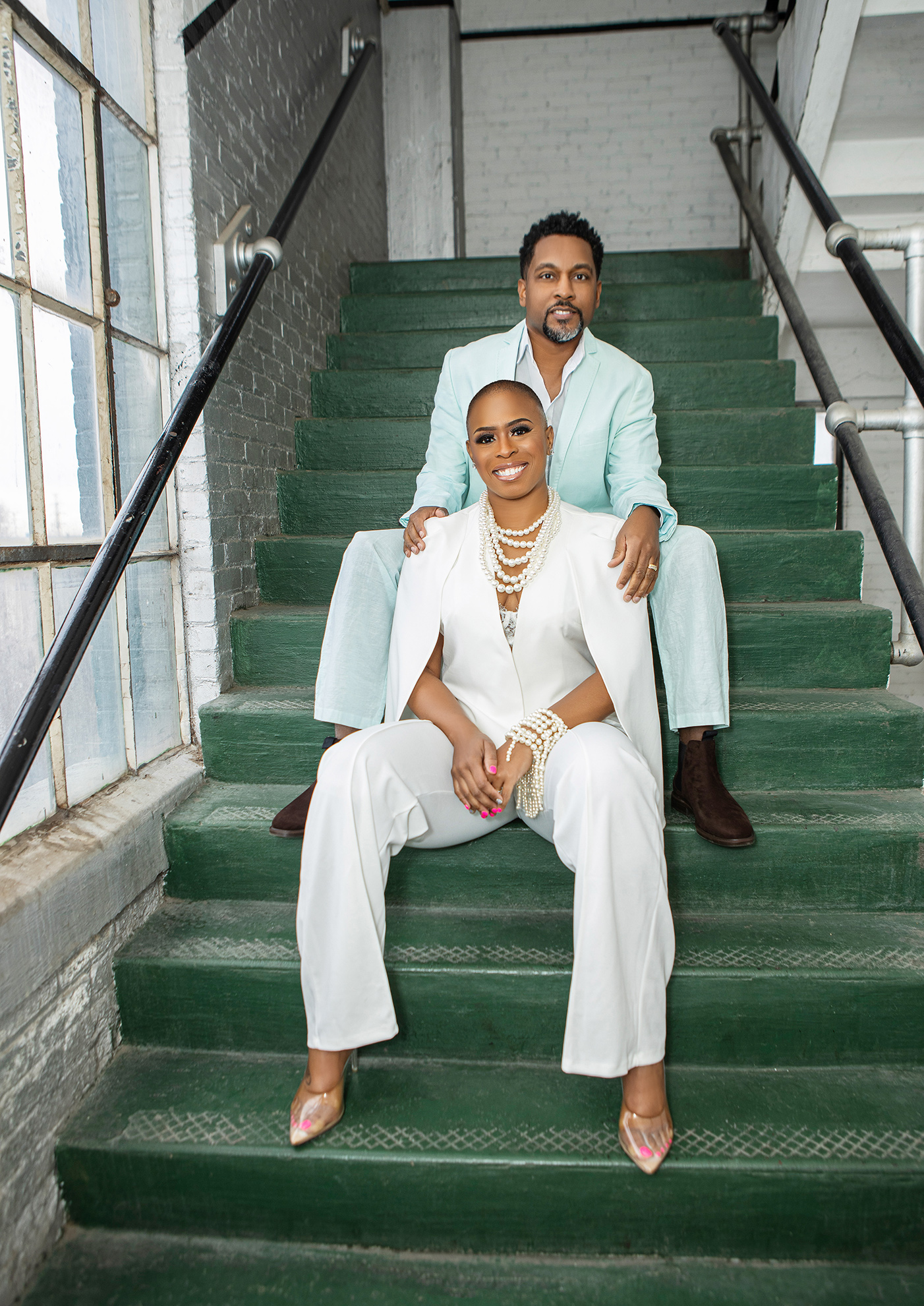 Eric and Phillitia Charlton on green stairs — Phillitia in a white suit with pearls seated in front, Eric in a mint blazer standing behind
