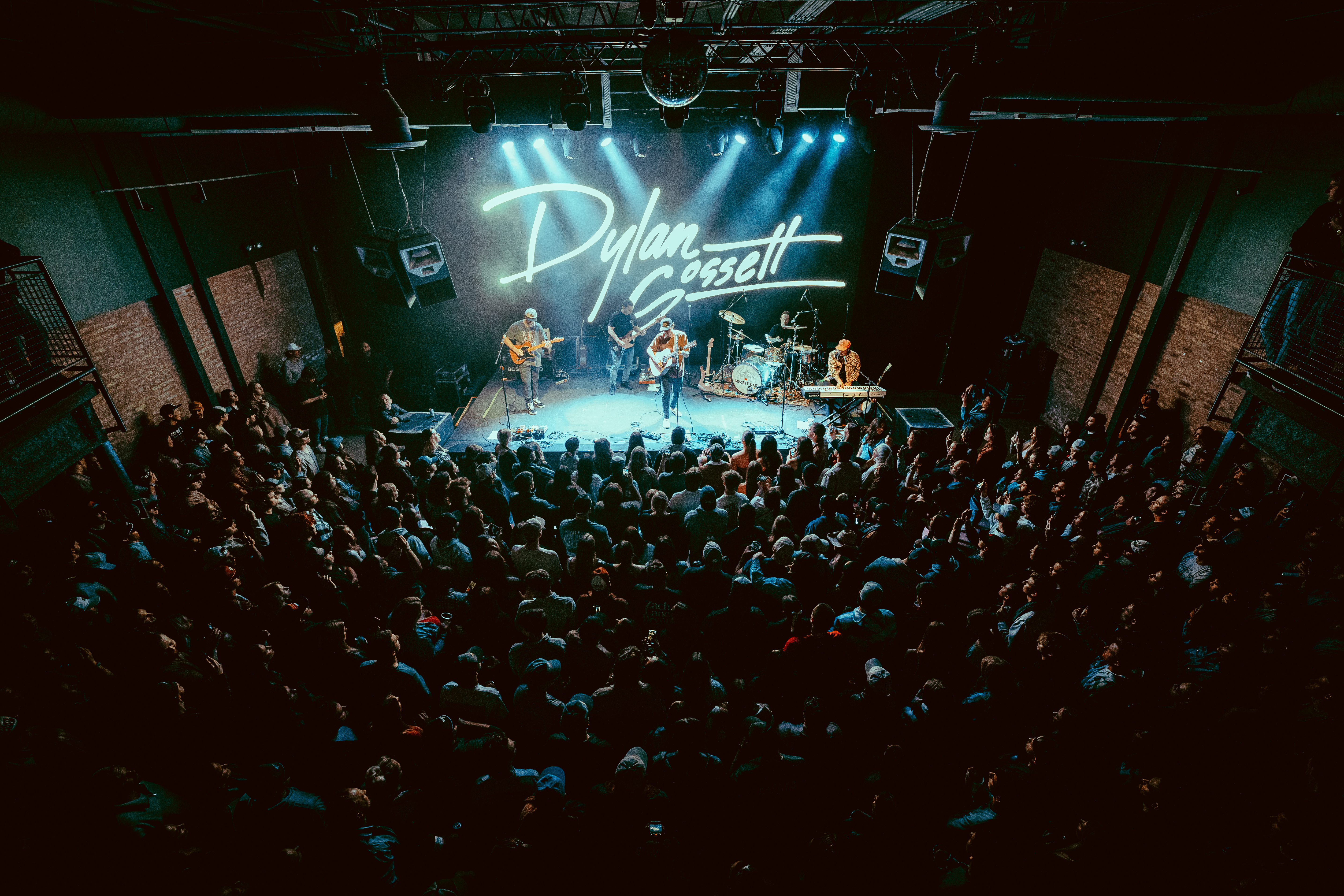 Overhead view of a packed crowd facing a stage with a glowing 'Dylan Gossett' neon sign, band performing under blue spotlights