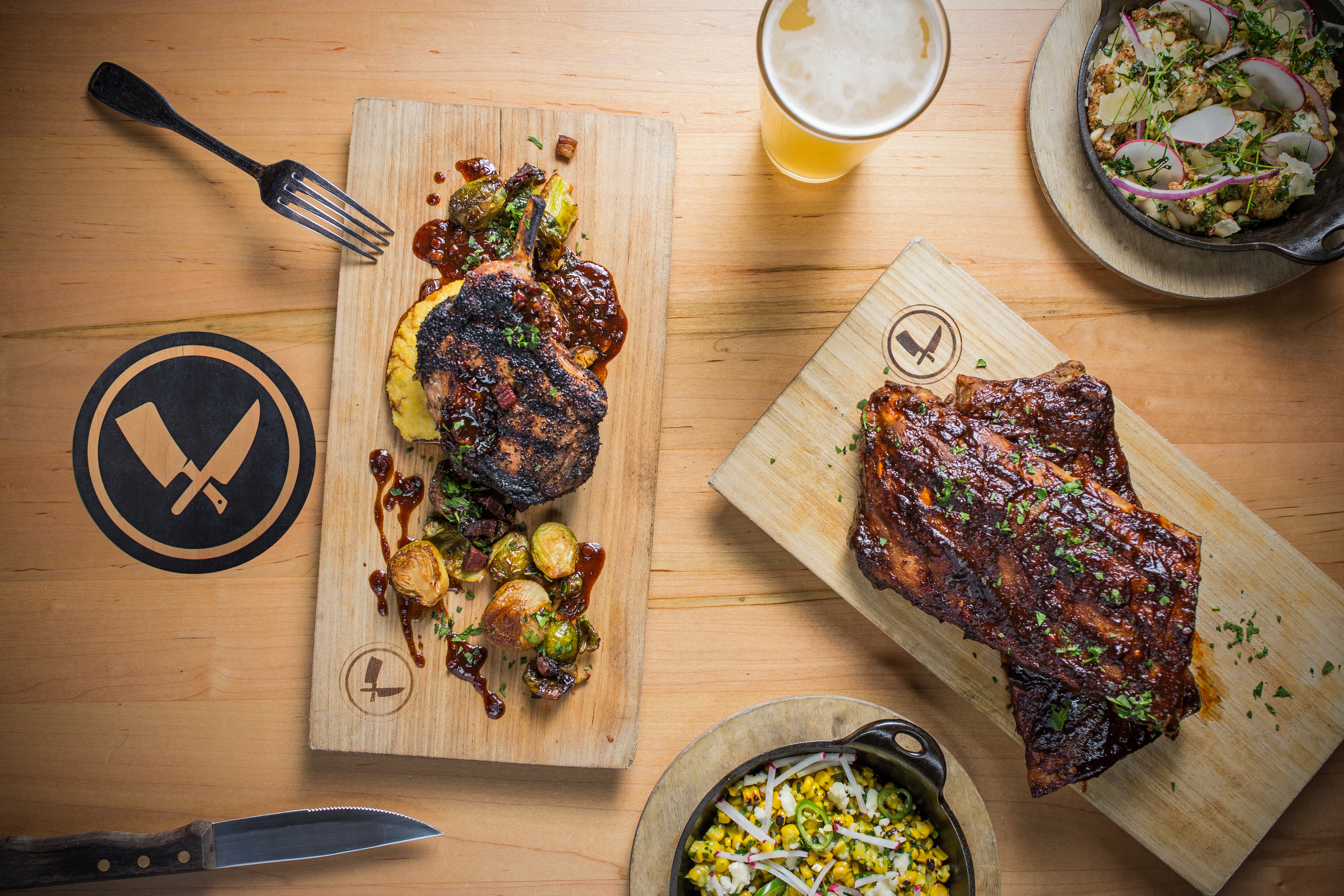 Overhead flat-lay of Chop Shop food: BBQ ribs on cutting boards, brussels sprouts, corn salad and a beer on a butcher-block table