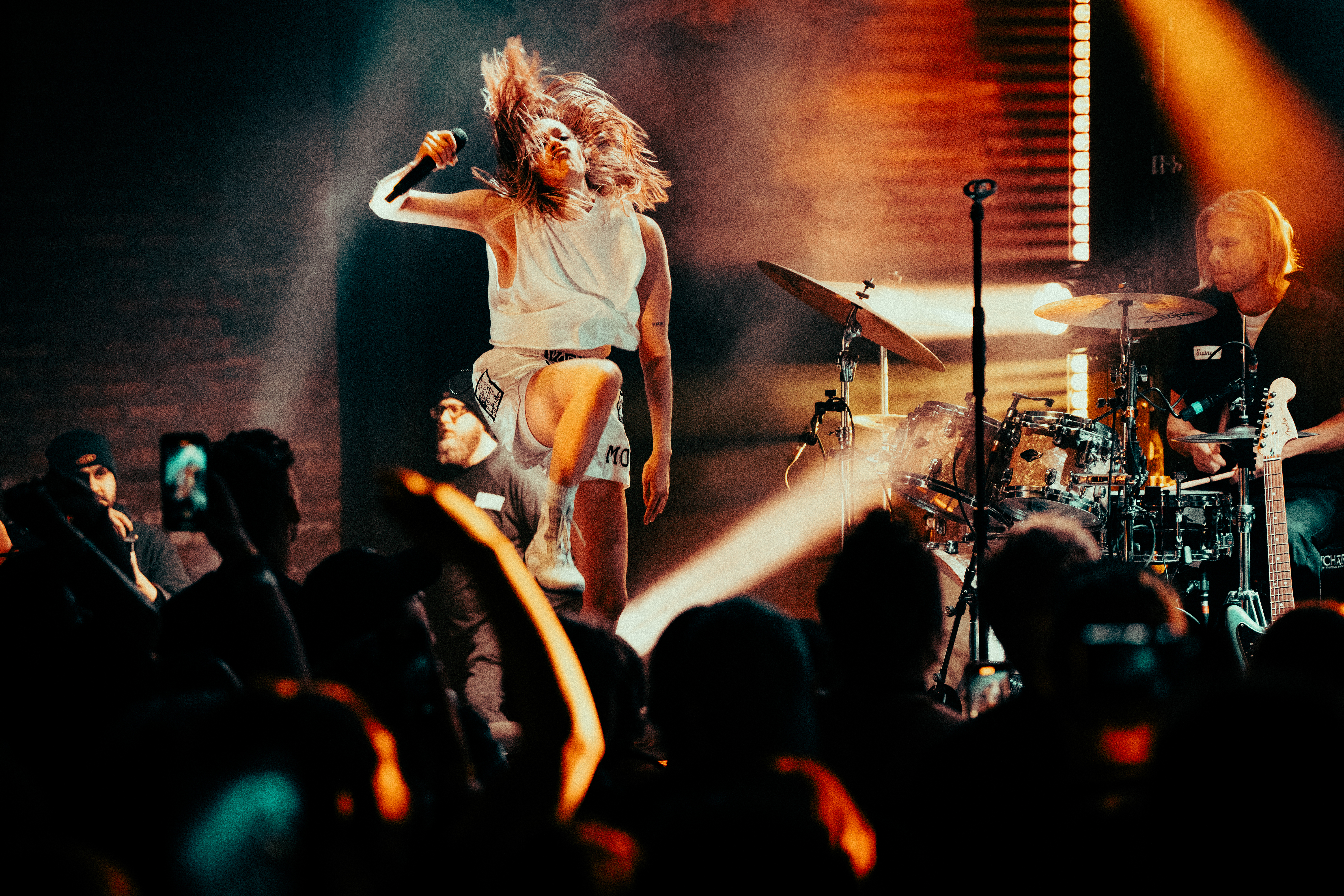 K.Flay performing with hair flying and a drummer behind her under orange stage light