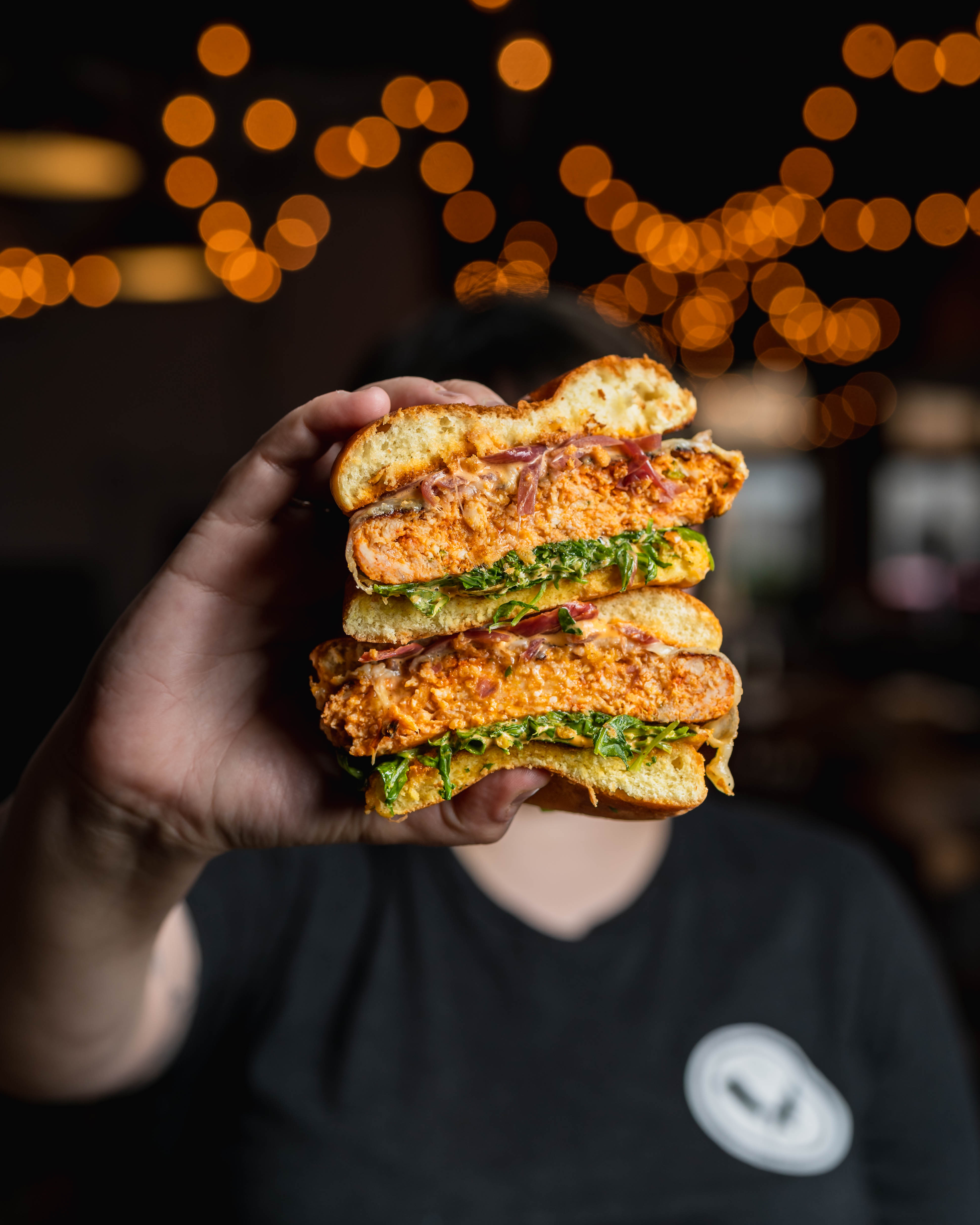 Hand holding up a stacked Chop Shop sandwich cross-section with string-light bokeh behind
