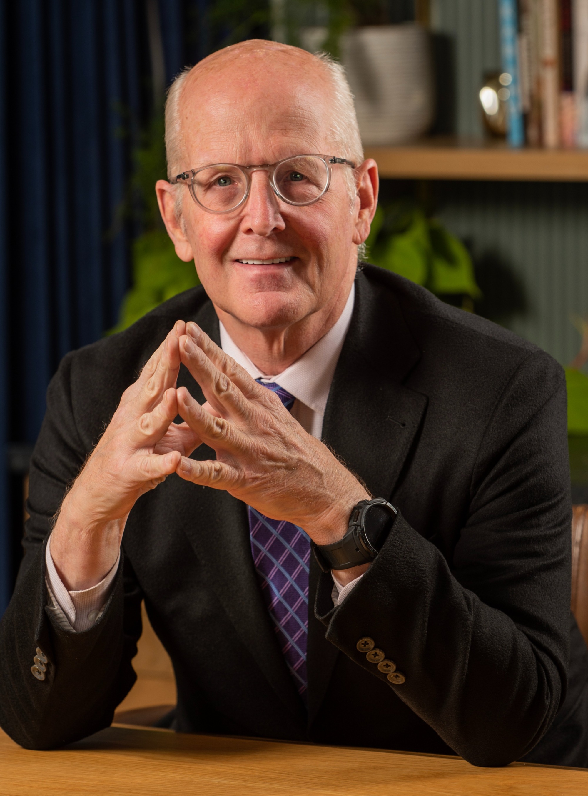 Liam Chrismer, older man with glasses in a dark suit and purple tie, hands steepled at a desk