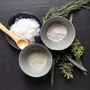 Overhead flat-lay of three bowls of spa scrub powders with rosemary and eucalyptus