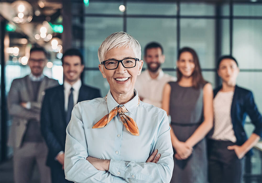 Confident leader with short white hair and glasses, arms crossed, standing with her diverse team in an office corridor