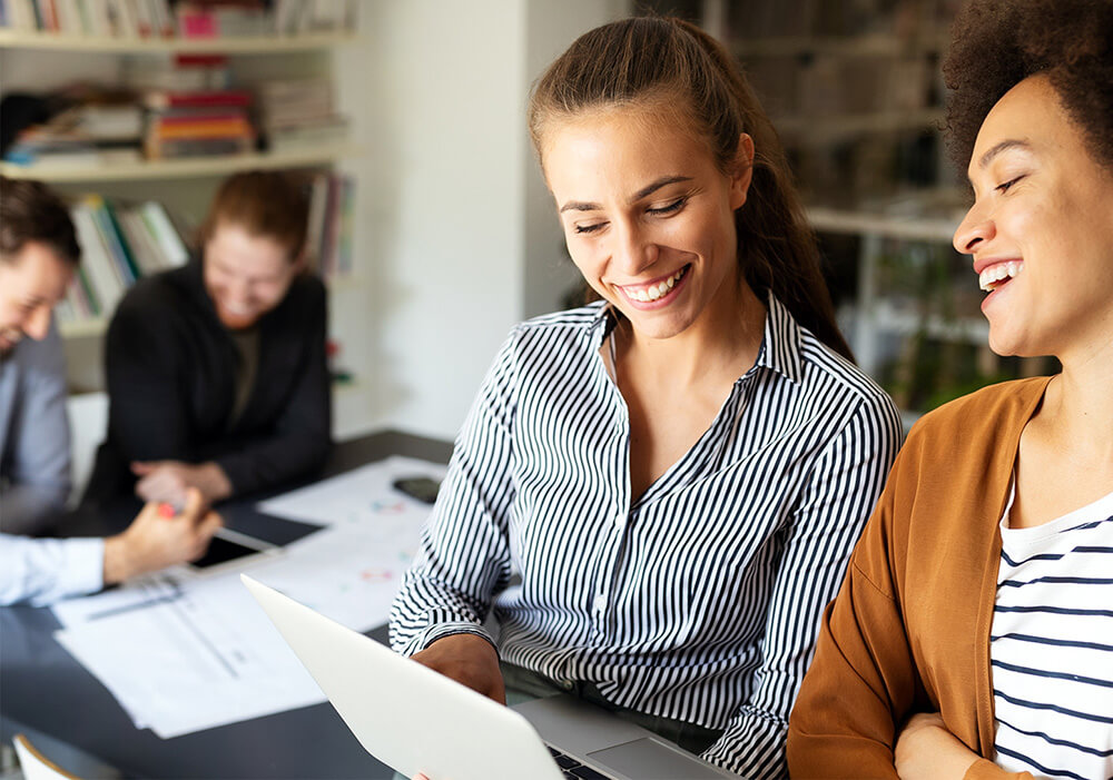 Two smiling women collaborating over a laptop in a bright office with colleagues in the background