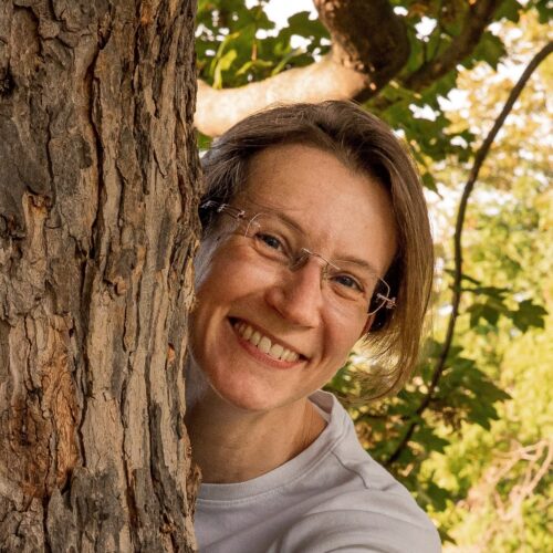 Christiane Helbig, Leadership Consultant & Coach — woman with short brown hair and wire-rimmed glasses smiling beside a tree trunk