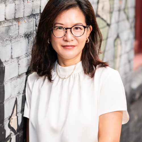 Wen Reese, Leadership Consultant & Coach — woman with dark hair and round glasses in a white top against a graffiti brick wall