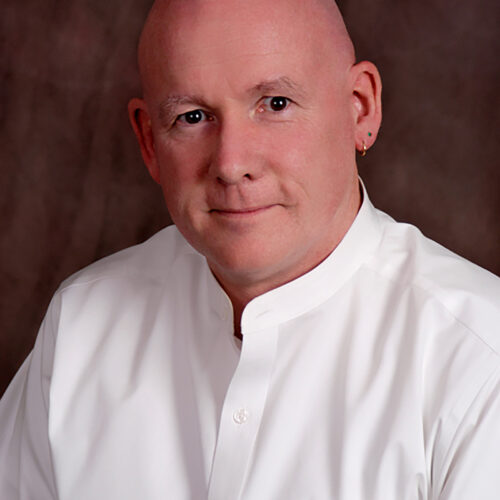 Dan Leahy, MA, Leadership Consultant & Coach — bald man in a white mandarin-collar shirt, studio portrait on a brown background
