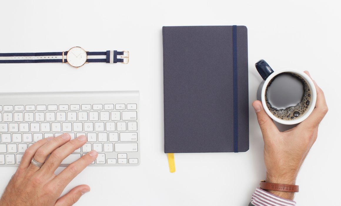 Overhead flat-lay of hands on a keyboard beside a notebook and coffee