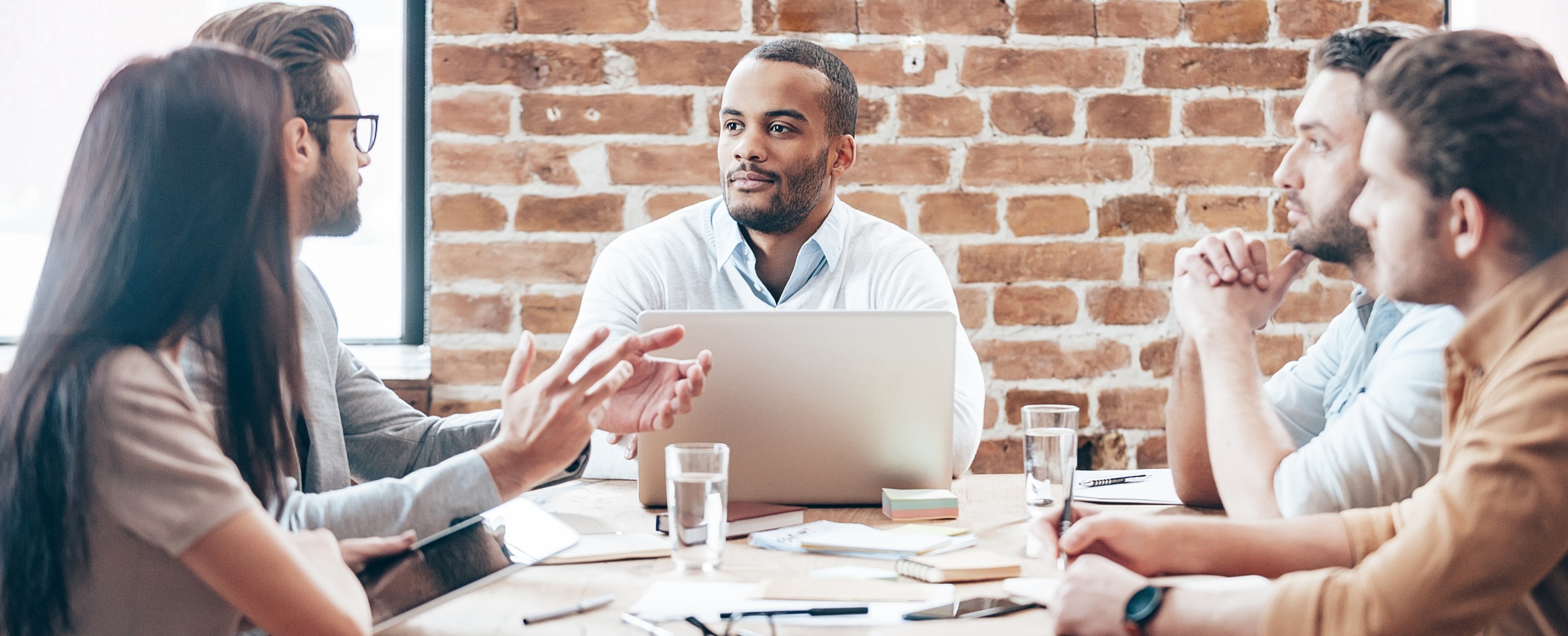 Five professionals in a team meeting around a table in a brick-walled office