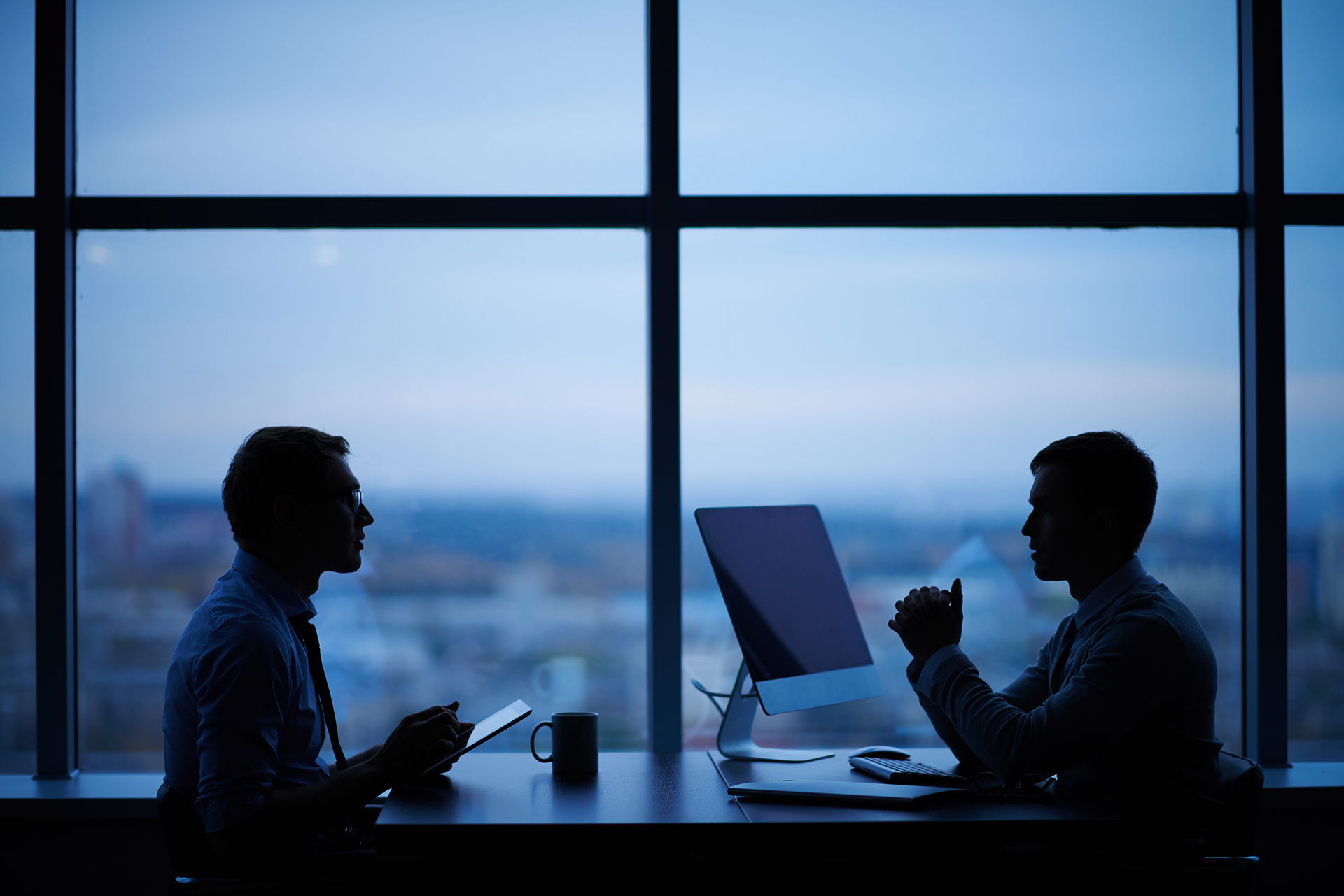 Silhouette of two men in a one-on-one meeting at a desk against floor-to-ceiling windows at dusk