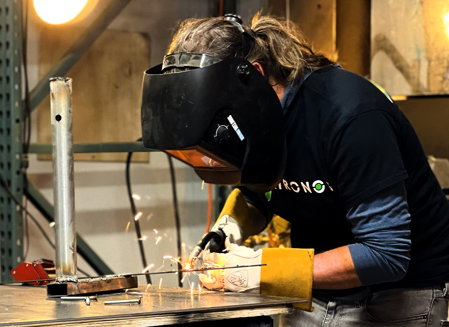 Person in a welding helmet and gloves welding metal with sparks flying at a workbench in the makerspace