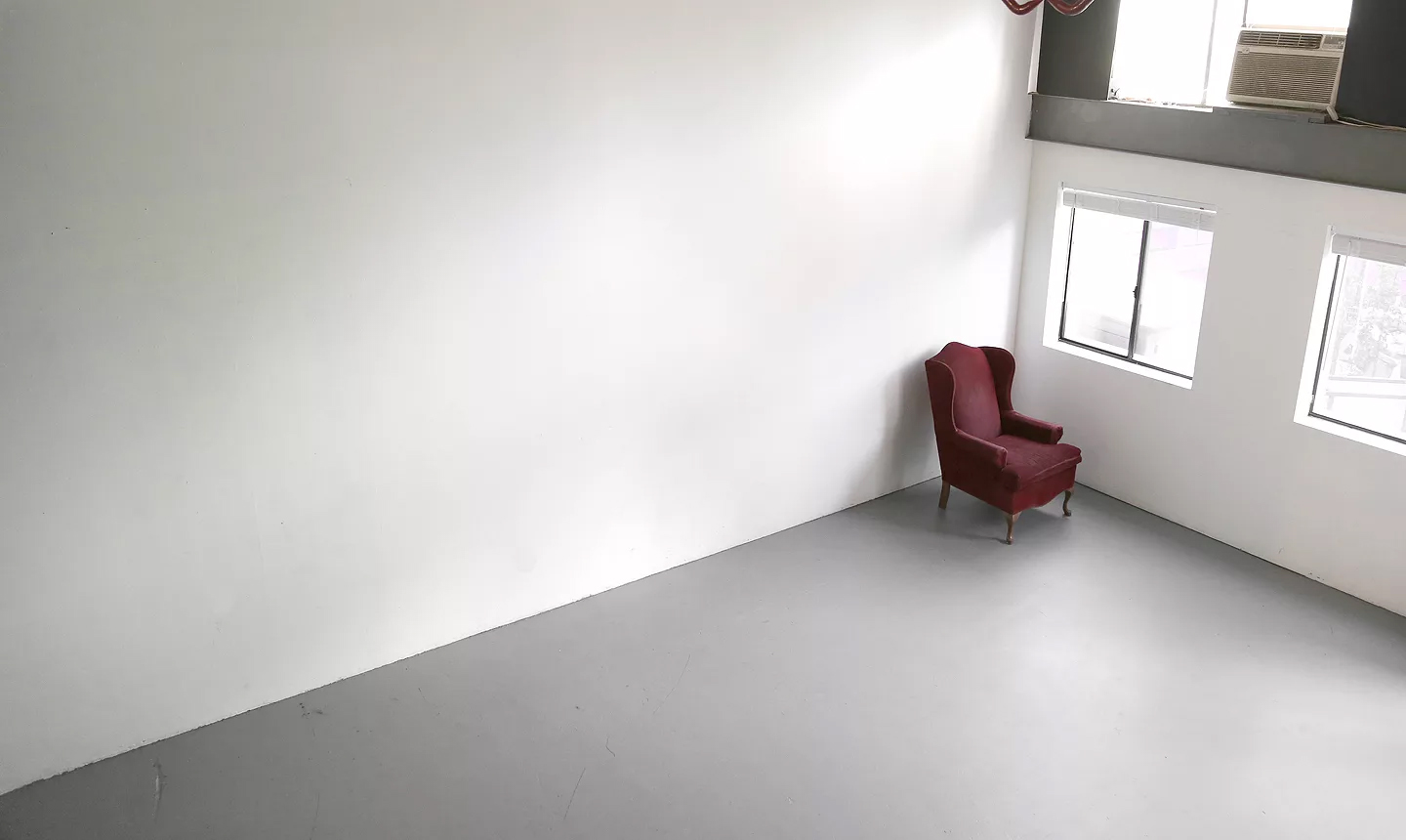 White-walled photo studio with grey floor and a single red wingback armchair by the windows