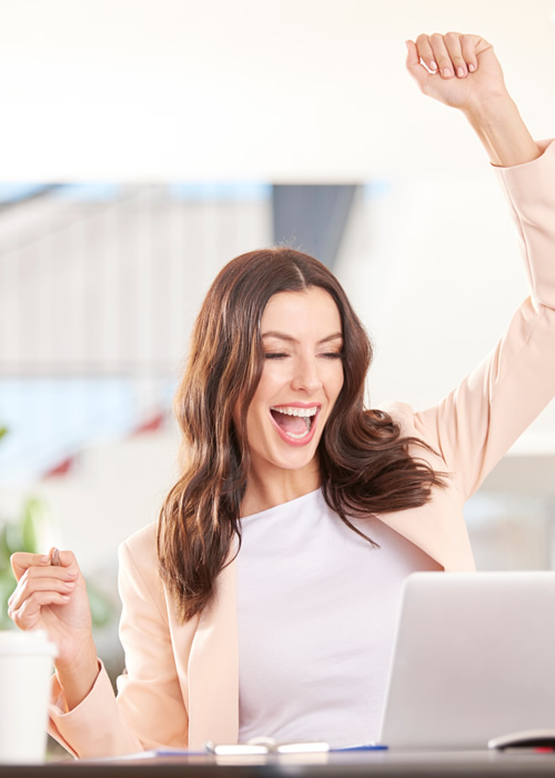 Woman in a pink blazer celebrating with raised arms in front of a laptop