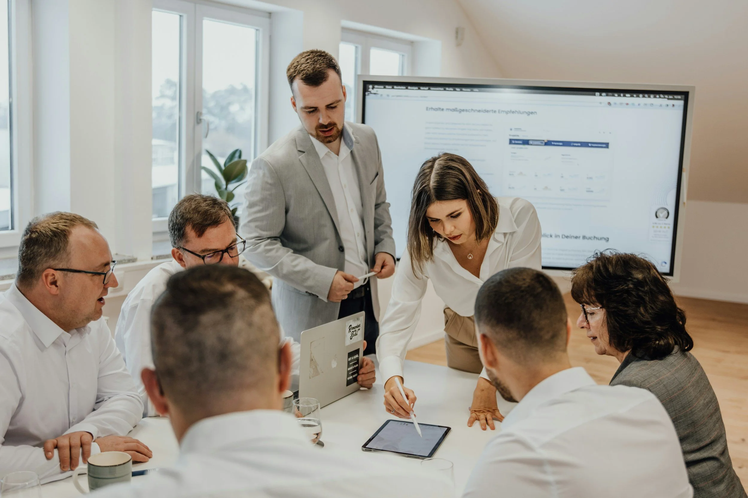 Six business professionals gathered around a white table collaborating, with a large presentation screen behind them.