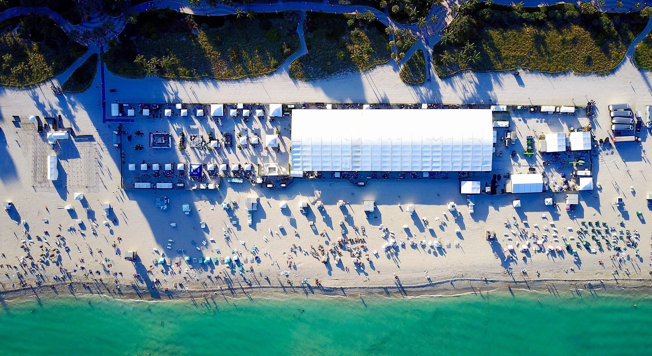 Aerial drone view of a large outdoor CREaM culinary event on Miami Beach with white tents beside turquoise ocean