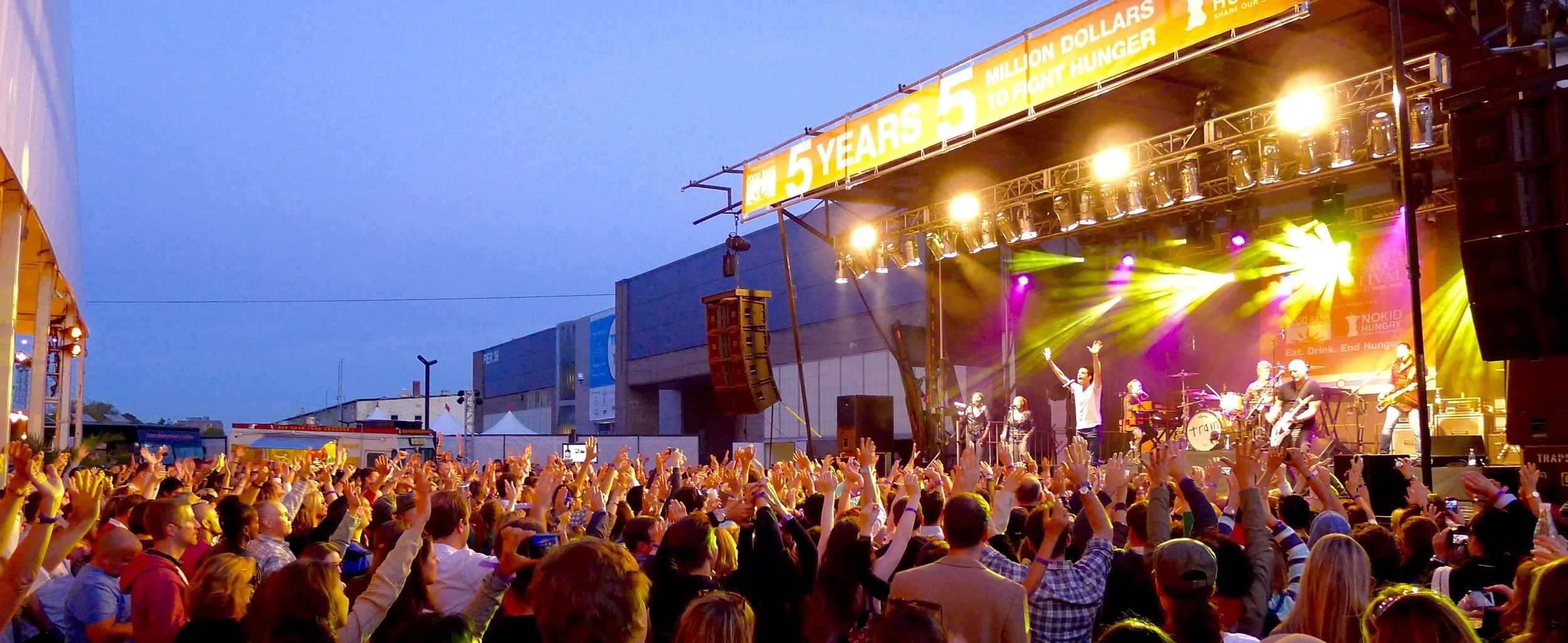 Outdoor concert crowd with hands raised toward a lit stage at dusk, banner reads 5 Years 5 Million Dollars to Fight Hunger