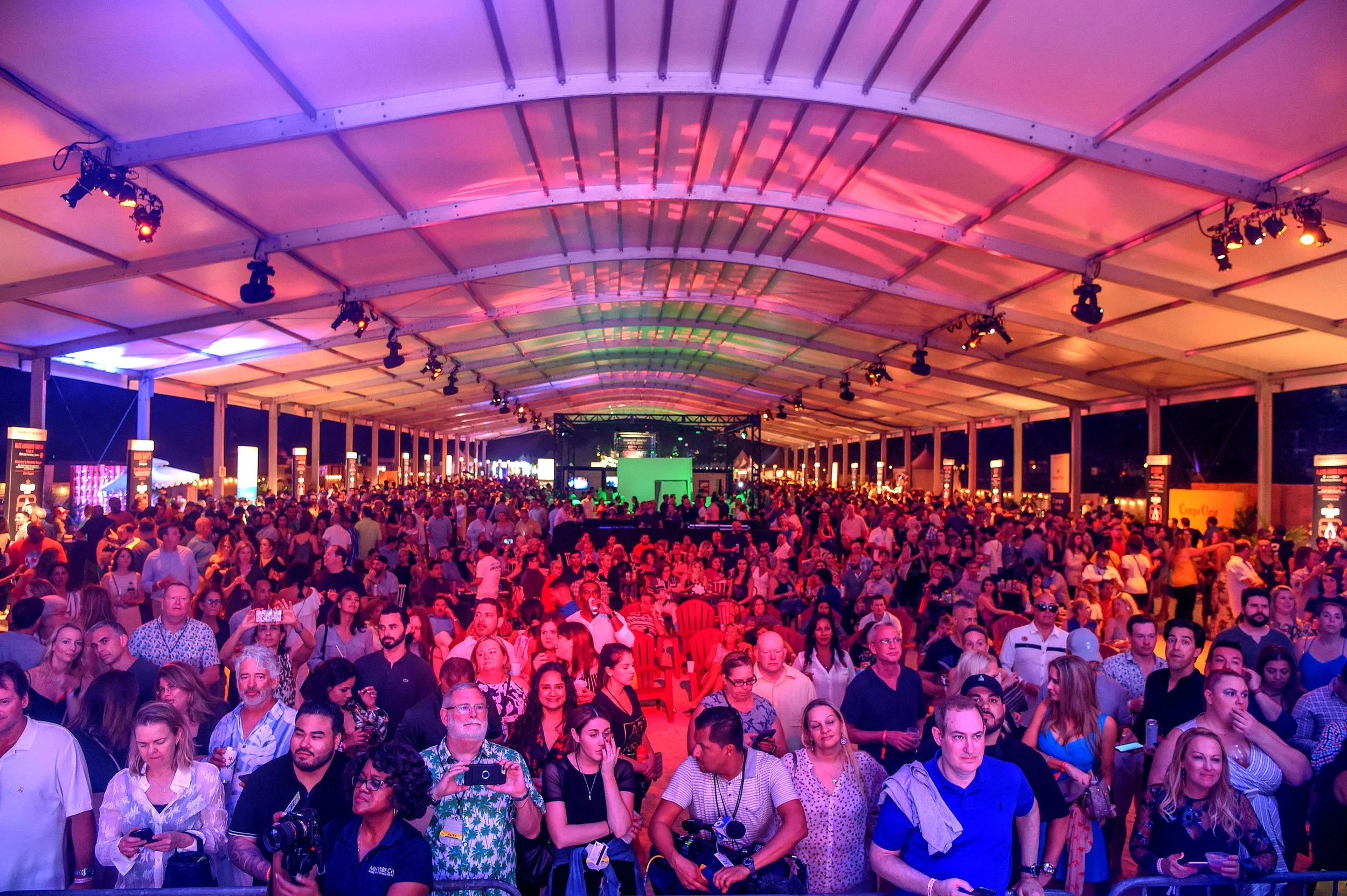 Large crowd of attendees under a lit tent at a culinary festival with pink and green stage lighting