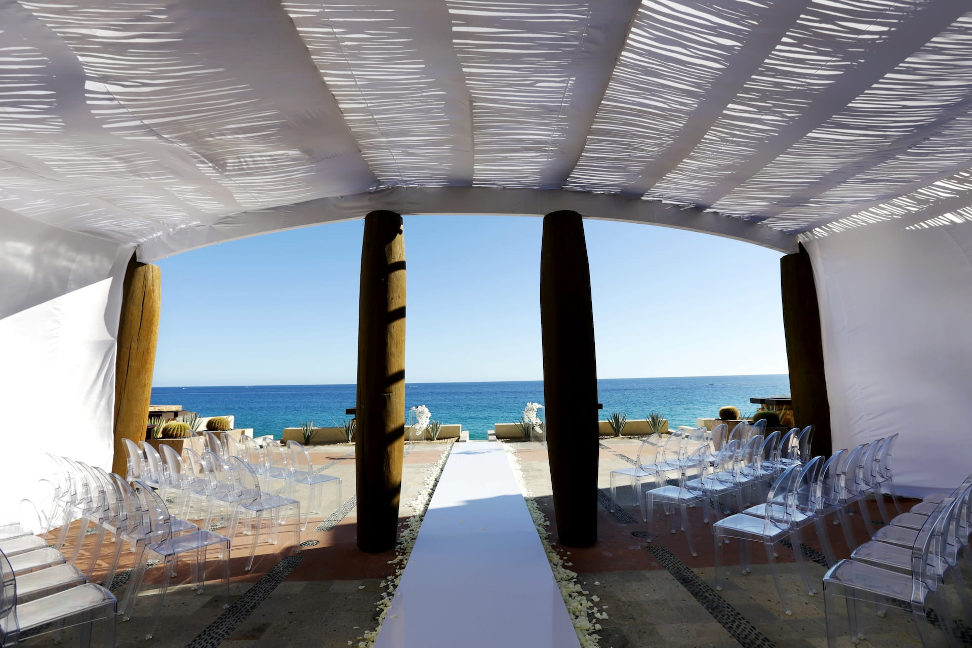 Open-air beachside ceremony aisle with acrylic ghost chairs under a white fabric canopy