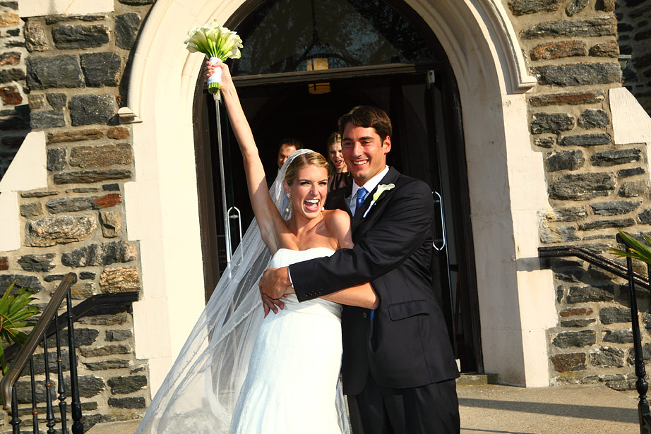Jubilant bride and groom exiting a stone church, bride holding her bouquet aloft