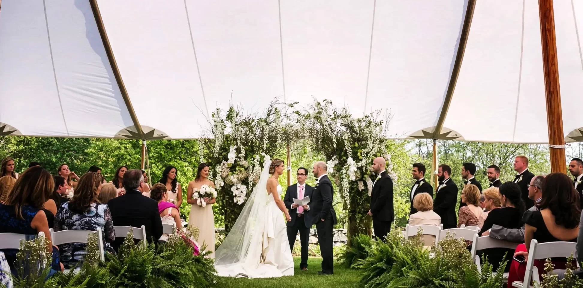 Wedding ceremony under a white sailcloth tent with the couple at a floral arch surrounded by the bridal party