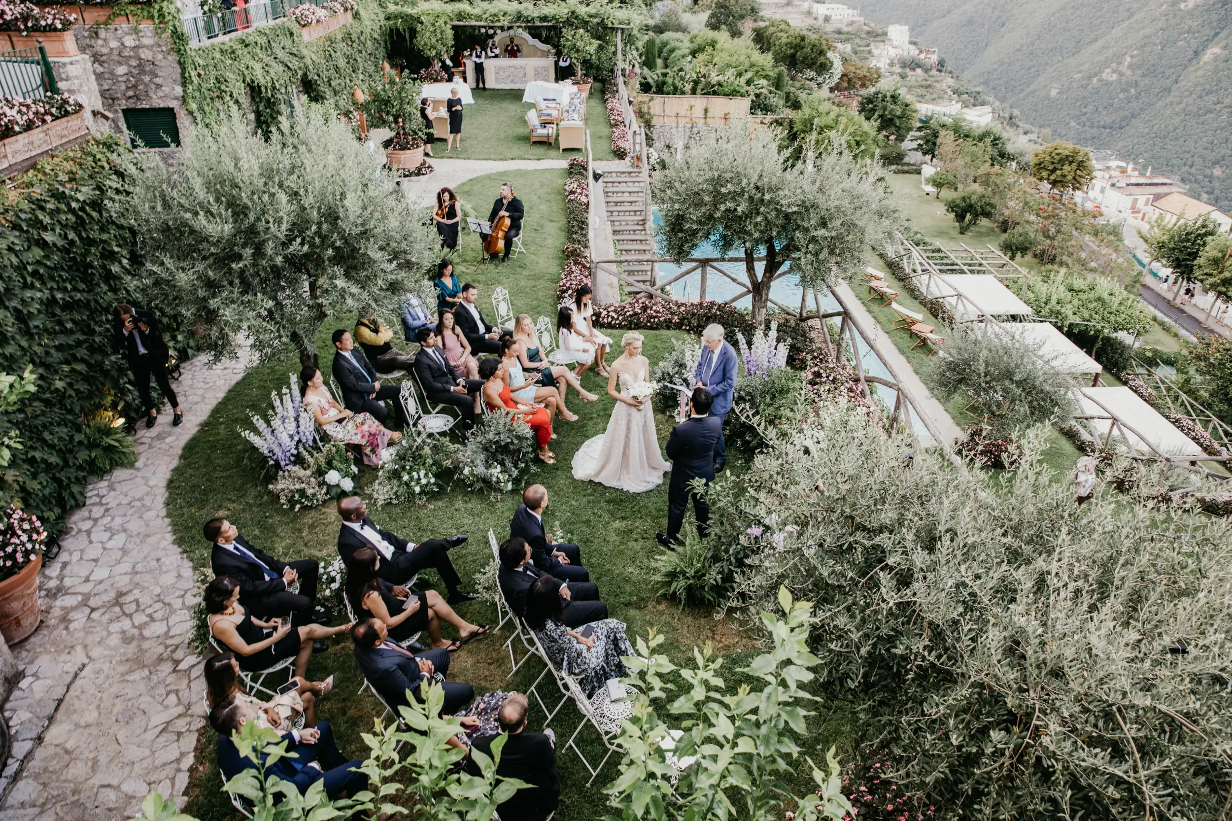 Aerial view of an outdoor Italian wedding ceremony in a lush terraced garden with a mountain valley backdrop