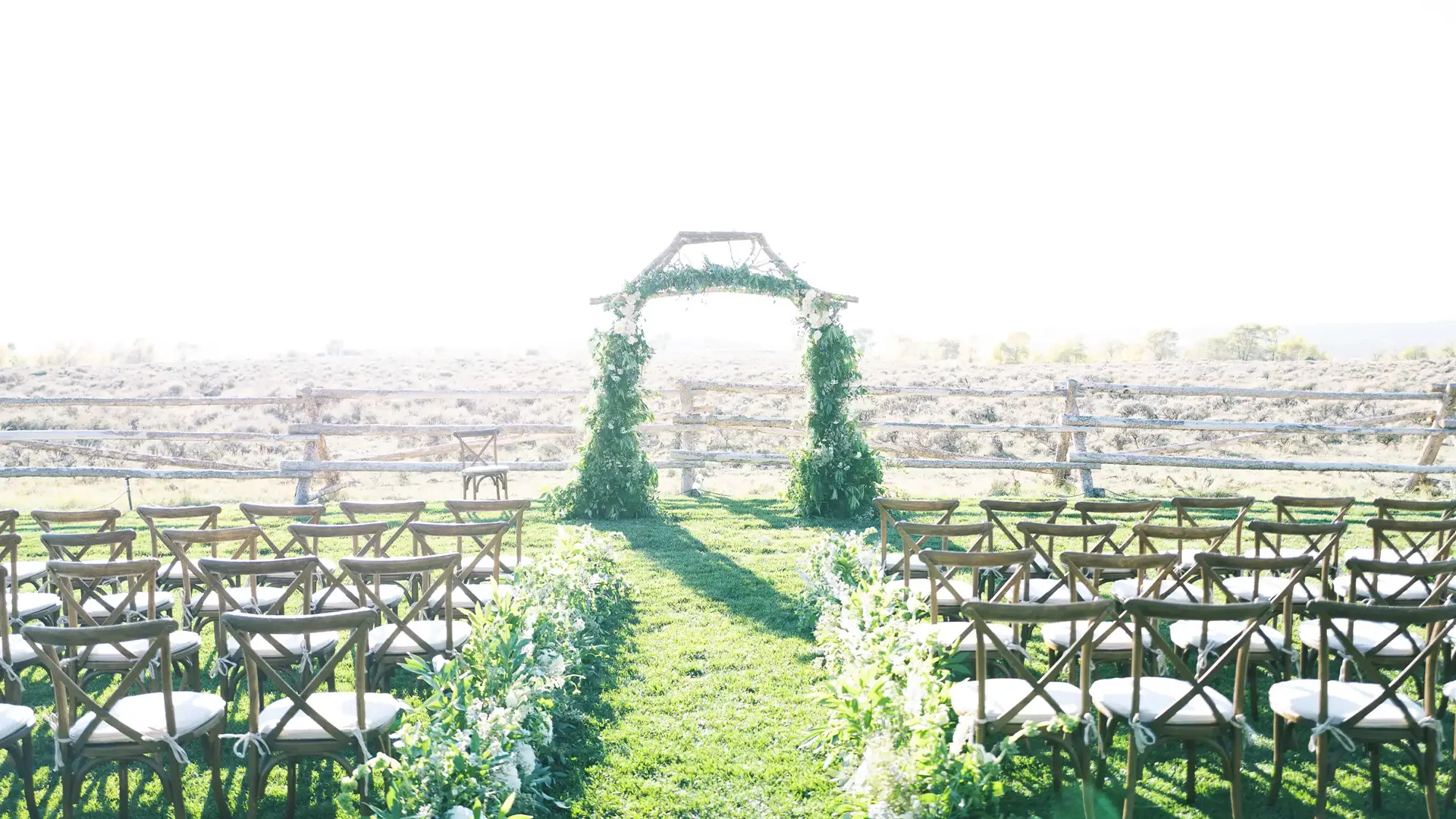 Bright outdoor ceremony with wooden cross-back chairs and a floral arch at the altar