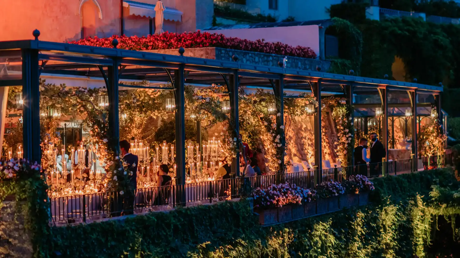 Glowing glass-enclosed event terrace adorned with candles and flowers at an Italian coastal venue at night