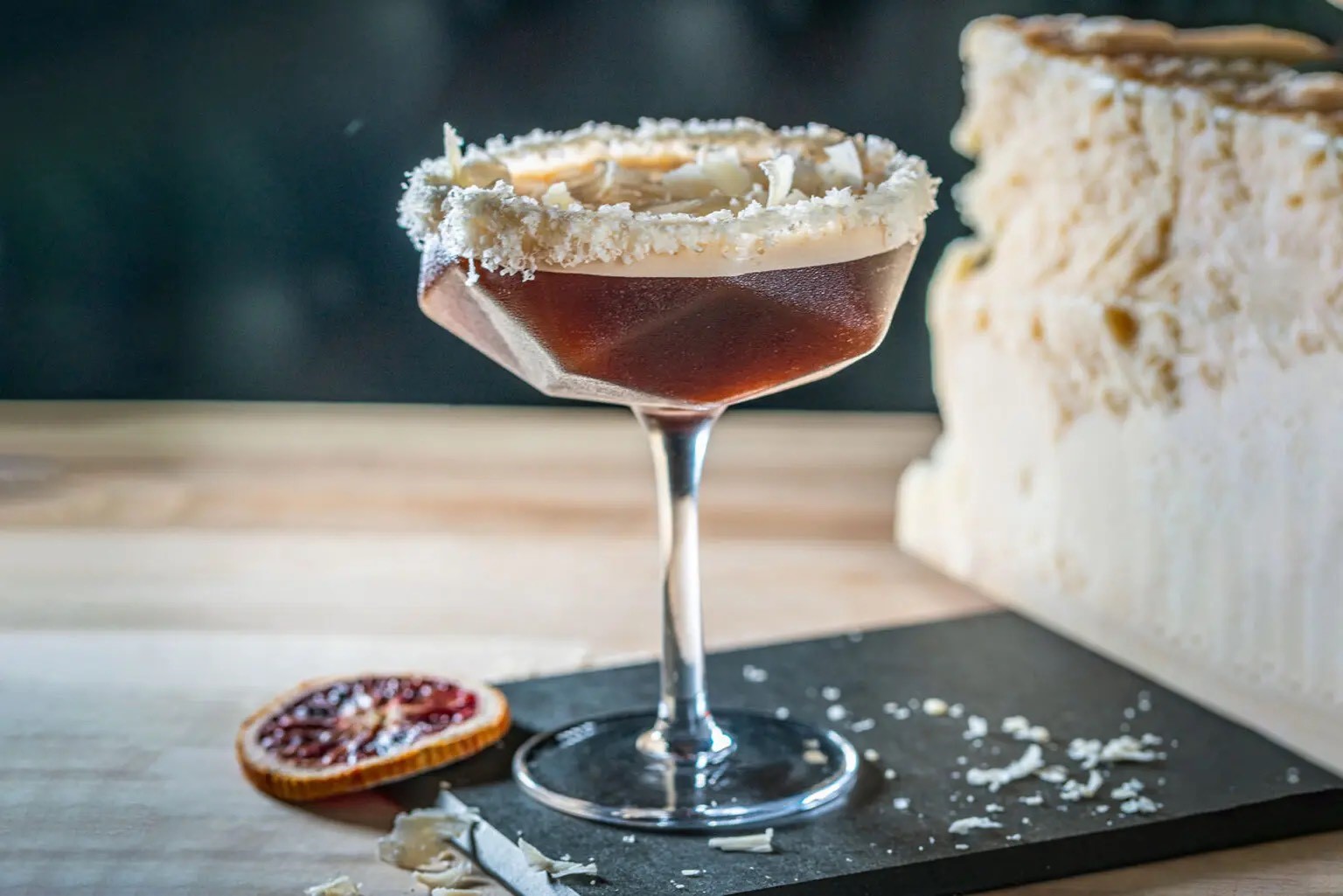 Dark cocktail in a faceted coupe with coconut rim and blood orange garnish