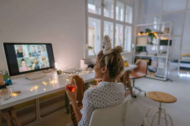 A guest raising a drink during a virtual reception on a desktop video call, festive lights behind