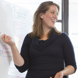 Woman in a black top smiling while presenting at a whiteboard