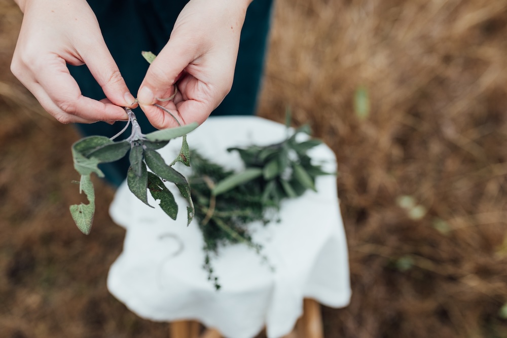 Hands bundling fresh sage and herbs outdoors in a dry field