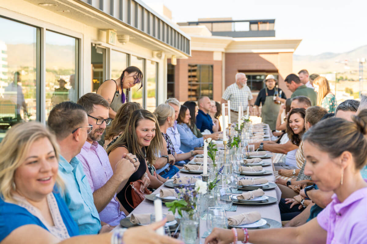 A large group at a long dinner table on a rooftop patio at sunset with the Boise hills behind